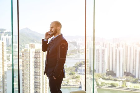 Male CEO  is standing in his office near window with view of business district background with copy space