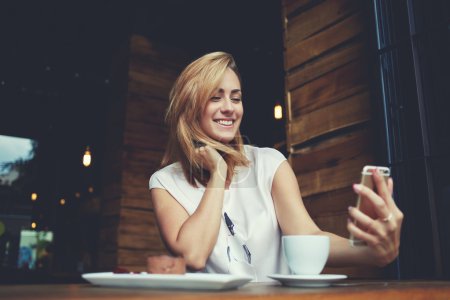 Attractive smiling hipster girl making self portrait on cell telephone while sitting in cafe 