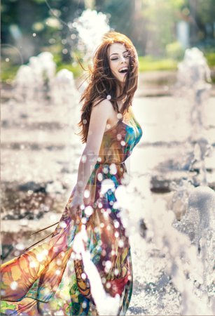 Attractive girl in multicolored short dress playing with water in a summer hottest day. Girl with wet dress enjoying fountains. Young beautiful happy female playing with outdoor water fountains.