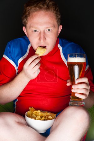 Close-up portrait of young man wearing sportswear fan of football team is watching tv and rooting for his favorite team. Sitting on beanbag alone at night drinking beer and eating chips