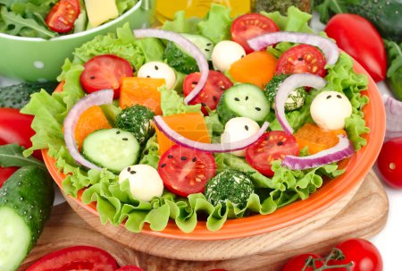 The Greek salad with cheese balls on an orange plate on a red checkered napkin and fresh ripe vegetables and herbs on a white background with a place for the text.