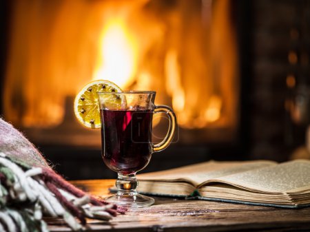 Hot mulled wine and a book on the wooden table. Fireplace with w