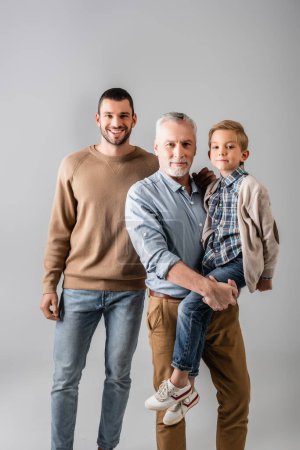 happy mature man holding grandson near cheerful son isolated on grey