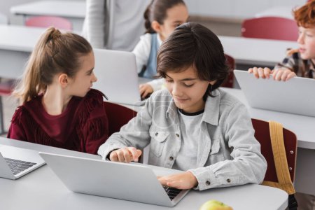 schoolboy typing on laptop in classroom near classmates on blurred background