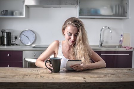 Blonde girl sitting at a kitchen counter using a tablet and holding a mug in her right hand