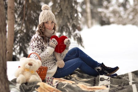 Attractive,beautiful,cute,pretty girl hold toy,girl with big,red heart in her hands,sit in warm plaid,has picnic in winter forest.Interesting,delightful,nice,attractive girl with big,read heart,toy in her hands in winter,snow,cold forest.