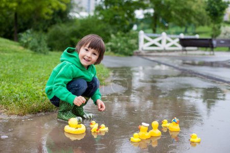 Little boy, jumping in muddy puddles in the park, rubber ducks i
