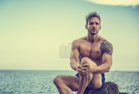 Handsome muscular man on the beach sitting on rocks