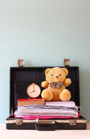 Open case with books towel teddy bear and vintage clock. vacation concept.