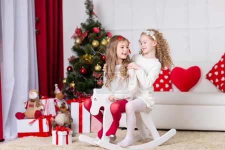 Christmas, x-mas, winter, happiness concept - two adorable happy girls playing on horse near the christmas tree