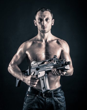 Confident young man shirtless portrait with machine gun against black background.