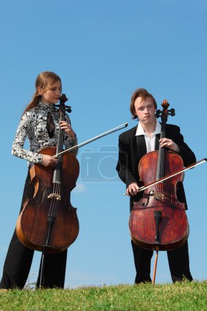 Two musicians play violoncellos against sky