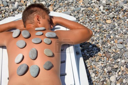 Young man sunbathes on beach with stones on back