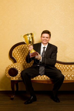 Smiling businessman with win cup in hand in suit sitting on sofa