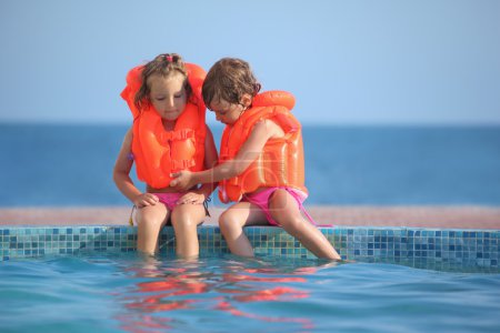 Two little girls in lifejackets sitting on ledge pool on resort