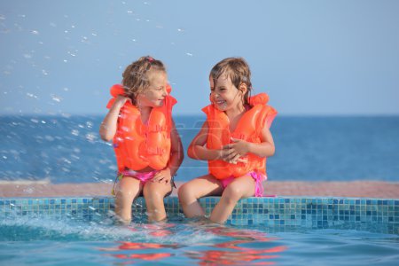 Two little girls in lifejackets sitting on ledge pool on resort,