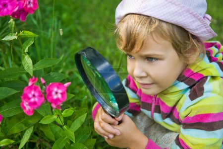 Pretty Little Girl with magnifying glass looks at flower
