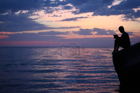 Silhouette guy sitting on breakwater in evening near sea, reads