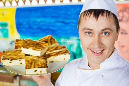 Cheerful cook in uniform holding cheese baked pudding on dish