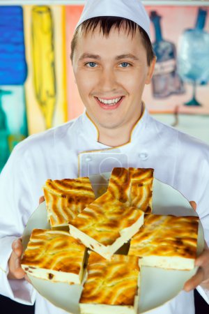 Cheerful cook in uniform holding cheese baked pudding on dish