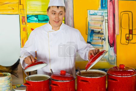 Cheerful cook in uniform near red pans in public catering restau