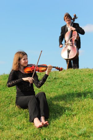 Violinist and violoncellist play on grass against sky