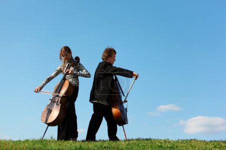 Two violoncellists play on grass against sky