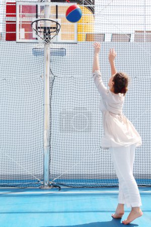 Brunette woman in white dress throws ball in basket, view from b