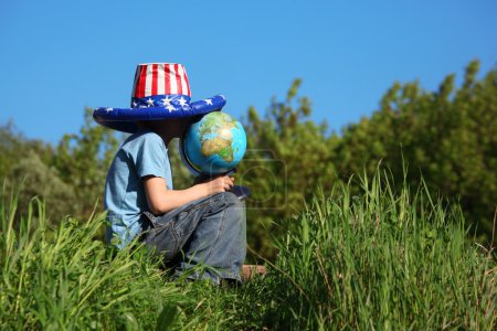Boy in big american flag hat sits on grass and holds globe