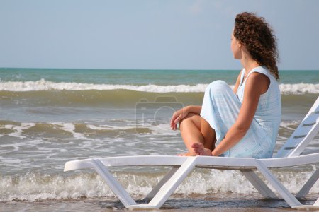 Young woman sits in the chaise lounge on the edge of the sea