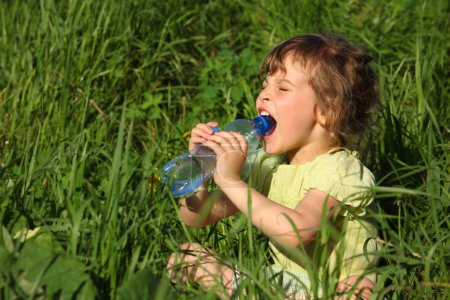 Girl sits in grass and drinks water from plastic bottle