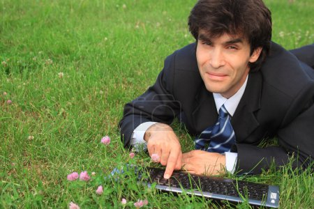 Smiling businessman lies on grass with laptop
