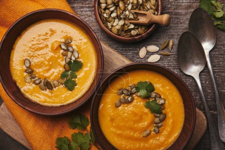 top view of bowls with tasty pumpkin soup, spoons and pumpkin seeds on wooden table