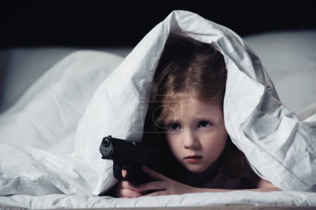 frightened child holding gun while hiding under blanket isolated on black
