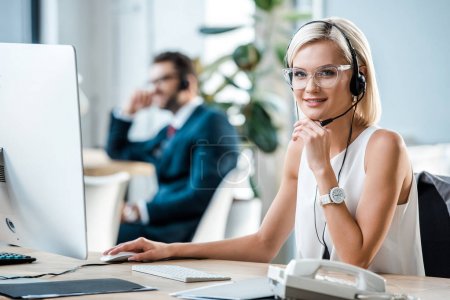 selective focus of cheerful blonde operator in headset looking at camera 