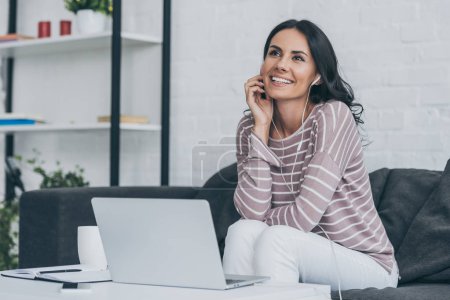 smiling woman looking away while sitting at table near laptop and listening music in earphones 