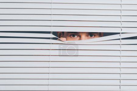 suspicious young man peeking and looking at camera through blinds, mistrust concept