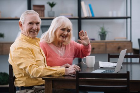 smiling senior couple sitting at table and using laptop during video call at home
