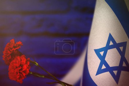 Israel flag with two red carnation flowers for honour of veterans or memorial day on blue blurred painted brick wall. Israel glory to the heroes of war concept.