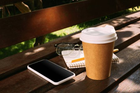 close up view of smartphone, notebook, eyeglasses and coffee to go on wooden bench