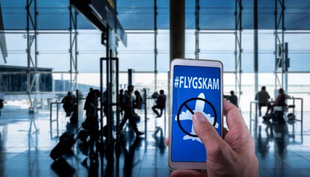 Hand holding a smartphone with Flyskam message on screen with airport passengers on the background.