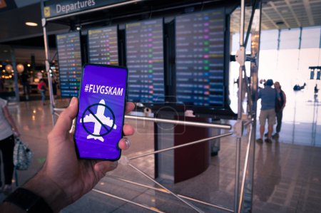 Hand holding a smartphone with Flyskam message on screen with airport timetables as background.