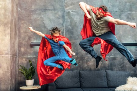 father and son in superhero capes and masks jumping on sofa at home