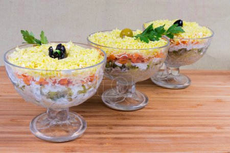 Salad made of canned fish, grated cheese, boiled eggs and vegetables, sprinkled with crumbled egg yolk called as mimosa salad in glass salad bowls on a wooden surface