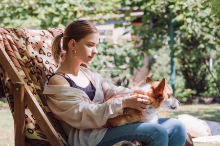 blonde girl holding welsh corgi dog on knees while sitting in deck chair in garden