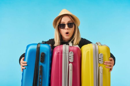 shocked girl in sunglasses and straw hat near multicolored travel bags isolated on blue