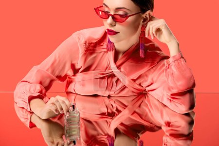 stylish woman in blouse and red sunglasses posing with perfume bottle and mirror reflection isolated on living coral