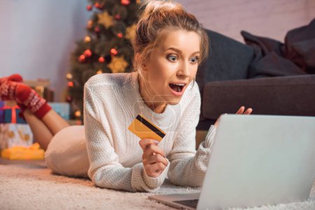 excited young blond woman lying on floor, holding credit card, using laptop and doing online shopping at christmas time 