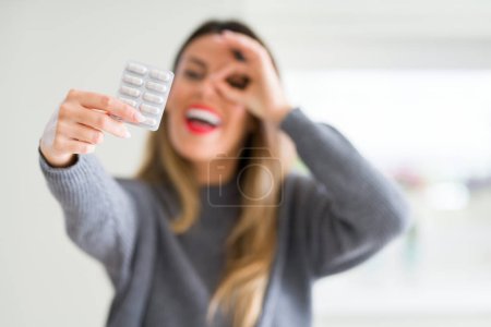 Young beautiful woman holding pharmaceutical pills at home with happy face smiling doing ok sign with hand on eye looking through fingers