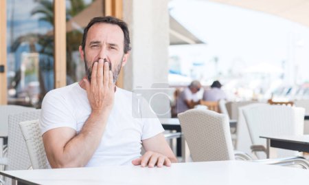 Handsome senior man sitting at terrace cover mouth with hand shocked with shame for mistake, expression of fear, scared in silence, secret concept